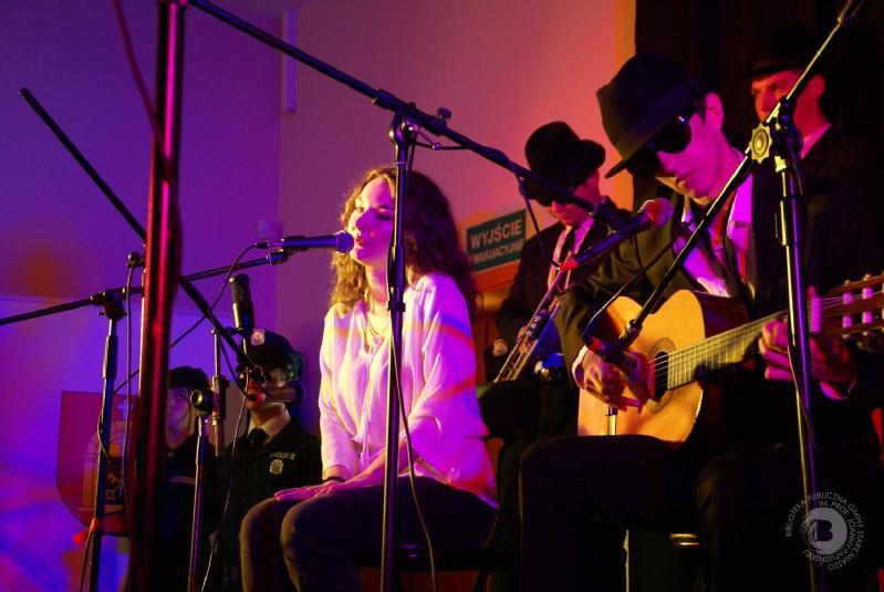 Kobieta śpiewa do mikrofonu na scenie, otoczona ludźmi w ciemnych garniturach, kapeluszach i okularach przeciwsłonecznych; jeden gra na gitarze akustycznej. Kolorowe oświetlenie tworzy żywą atmosferę.