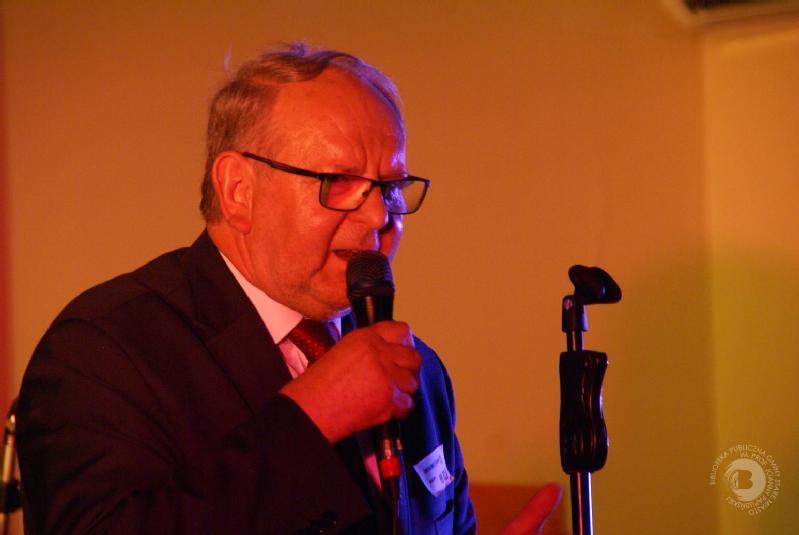 Starszy mężczyzna w garniturze i okularach mówi do mikrofonu w ciepłym oświetleniu, stojąc obok statywu mikrofonowego. Tło jest proste i delikatnie oświetlone.