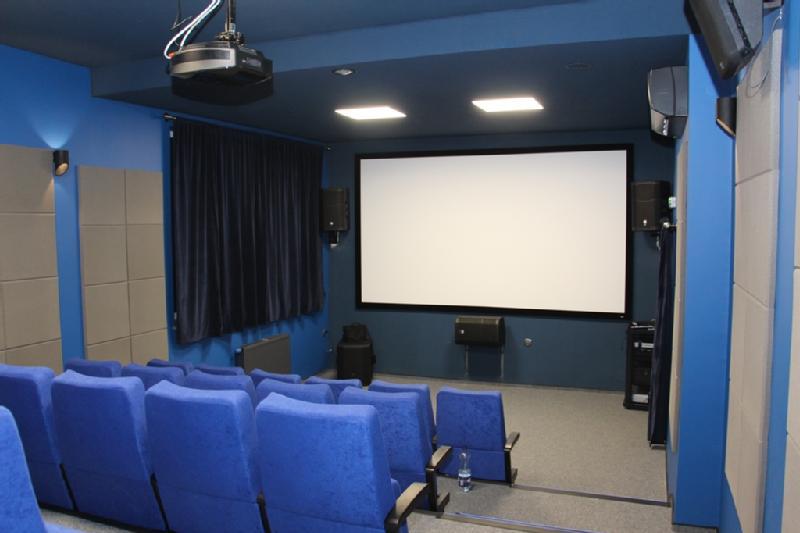 Mała, nowoczesna sala kinowa z niebieskimi ścianami i niebieskimi, wyściełanymi siedzeniami naprzeciwko dużego, białego ekranu projekcyjnego. W sali znajdują się głośniki, projektor montowany na suficie i panele pochłaniające dźwięk.