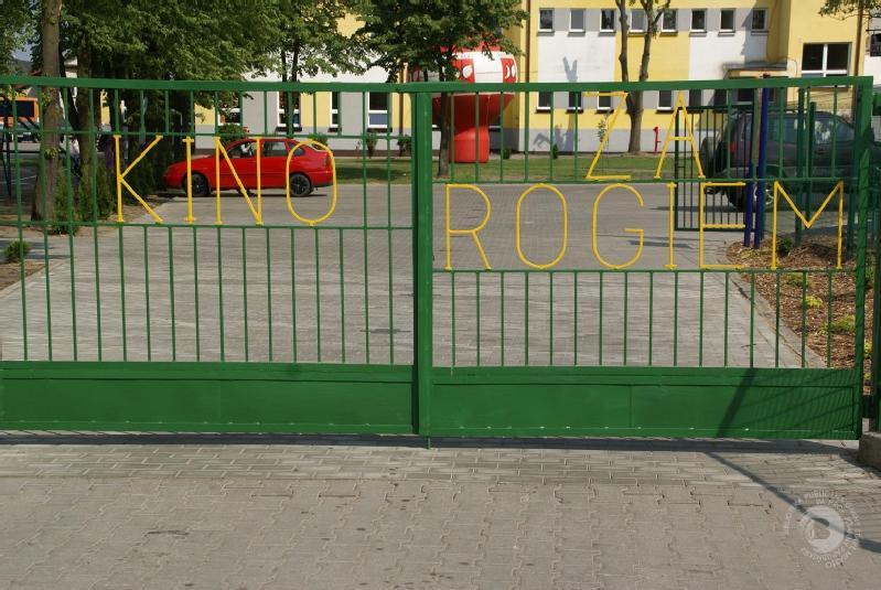 Zielona metalowa brama z żółtymi literami, na których widnieje napis KINO ZA ROGIEM; za bramą znajduje się czerwony samochód, drzewa i żółty budynek.