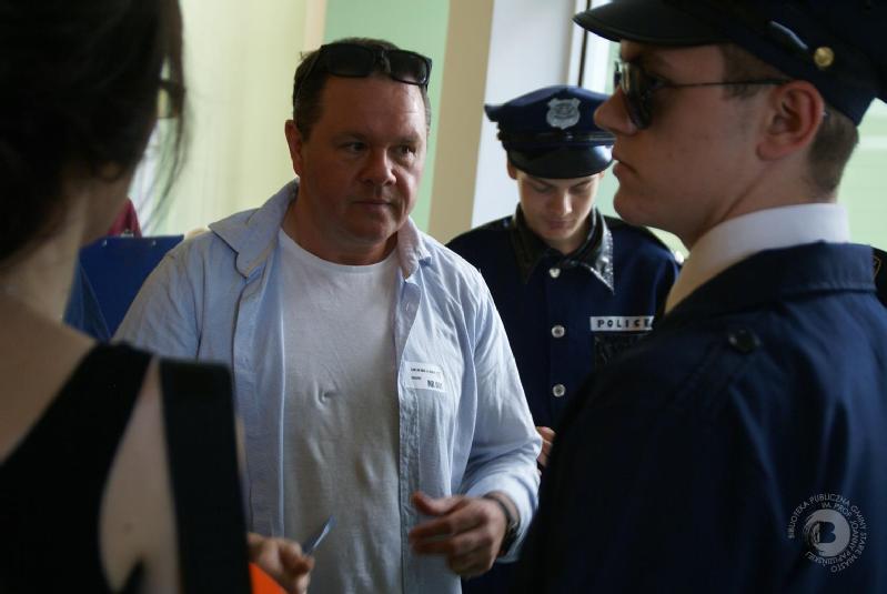 Mężczyzna w jasnoniebieskiej koszuli stoi w pomieszczeniu, trzymając długopis, zwrócony twarzą do osoby w mundurze policjanta. Inna osoba w stroju policjanta pisze w tle. Po lewej stronie częściowo widoczna jest kobieta.