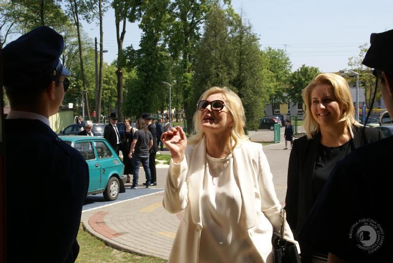 Kobieta w białym stroju i okularach przeciwsłonecznych uśmiecha się i gestykuluje, rozmawiając z umundurowanymi funkcjonariuszami na zewnątrz, obok niej stoi inna kobieta, a w tle widać kilka osób i samochodów.