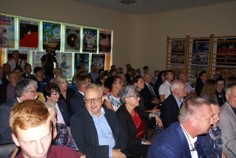 Grupa ludzi siedzi blisko siebie w dobrze oświetlonym pomieszczeniu, uważnie patrząc przed siebie. Na ścianie za nimi wiszą plakaty, a wszyscy wydają się zaangażowani, niektórzy rozmawiają, inni czekają na rozpoczęcie wydarzenia.