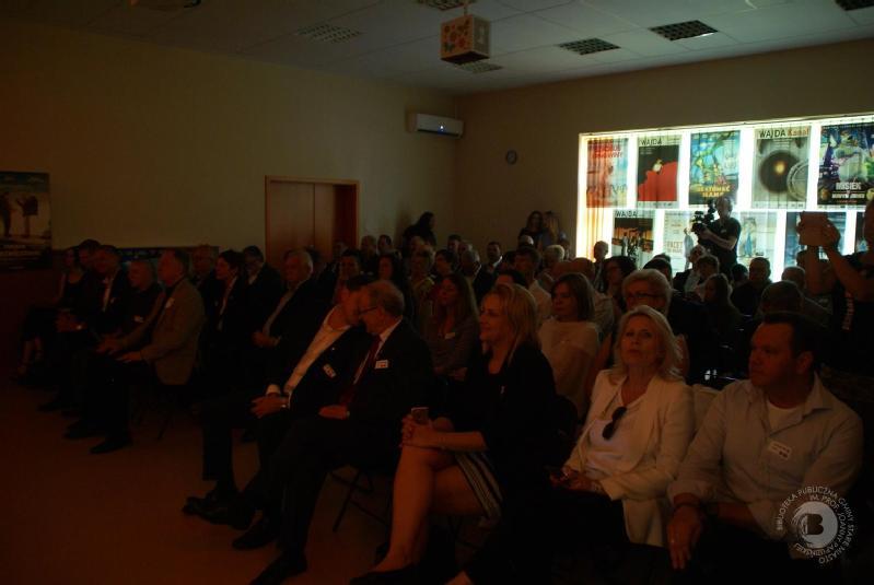 Grupa ludzi w formalnych strojach siedzi blisko siebie w słabo oświetlonym pomieszczeniu, uważnie patrząc w przód. Jasne plakaty i światło słoneczne są widoczne przez okna po prawej stronie pomieszczenia.