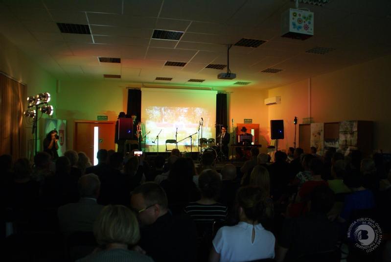 Publiczność siedzi w słabo oświetlonym pomieszczeniu i obserwuje scenę z mikrofonami i instrumentami, podczas gdy za wykonawcami na ekranie wyświetlany jest obraz przyrody.