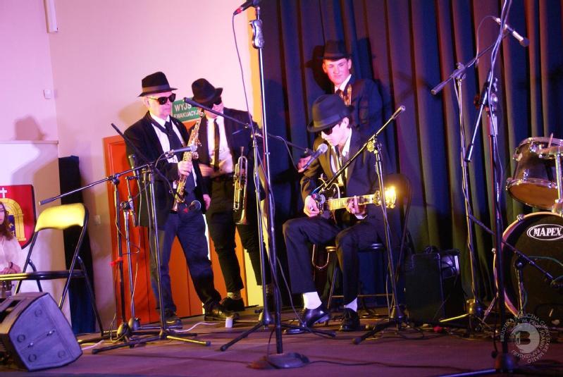 Zespół ubrany w czarne garnitury, kapelusze i okulary przeciwsłoneczne występuje na scenie z saksofonem, gitarą i innymi instrumentami. Scena jest oświetlona żółtymi i fioletowymi światłami, a po prawej stronie widoczny jest zestaw perkusyjny.