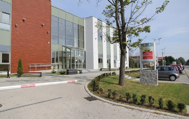 Nowoczesny budynek z dużymi szklanymi oknami i mieszanką drewna i białych paneli. Przed budynkiem znajduje się drzewo i przycięte krzewy, znak przy wejściu i parking z kilkoma samochodami obok budynku.