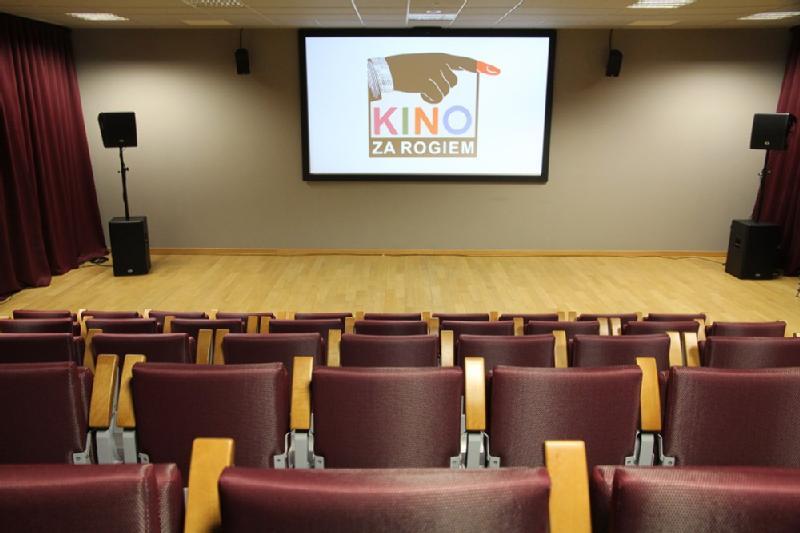 Małe kino z pustymi czerwonymi krzesłami zwróconymi w stronę ekranu wyświetlającego tekst „Kino Za Rogiem” i grafikę wskazującej ręki. W pokoju jest drewniana podłoga, beżowe ściany i bordowe zasłony po bokach.