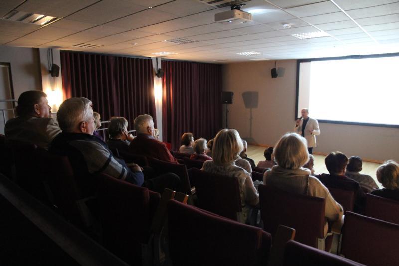 Grupa ludzi siedzi w ciemnym audytorium, obserwując osobę w jasnej kurtce wygłaszającą prezentację przed dużym, jasnym ekranem. Publiczność wydaje się być uważna, a pomieszczenie jest słabo oświetlone.