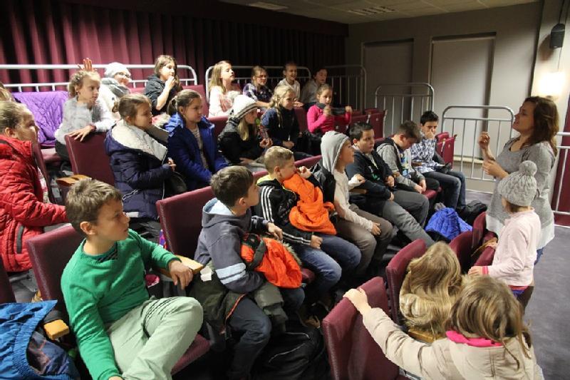 Grupa dzieci siedząca na piętrowych siedzeniach, słuchająca kobiety mówiącej z przodu sali. Niektóre dzieci mają podniesione ręce, a atmosfera wydaje się być edukacyjna i interaktywna.