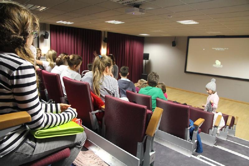 Grupa ludzi siedzi w małym teatrze, zwrócona twarzą do ekranu projekcyjnego z przodu. Niektórzy siedzą, podczas gdy inni dopiero wchodzą. W sali są czerwone siedzenia, bordowe zasłony i częściowo oświetlony sufit.