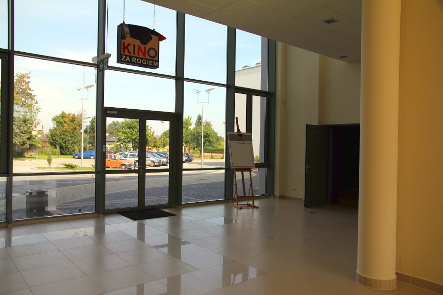 Przestronne lobby z dużymi oknami od podłogi do sufitu, kafelkową podłogą, szyldem Kino Za Rogiem i sztalugą przy otwartych drzwiach. Na zewnątrz zaparkowane samochody i drzewa.