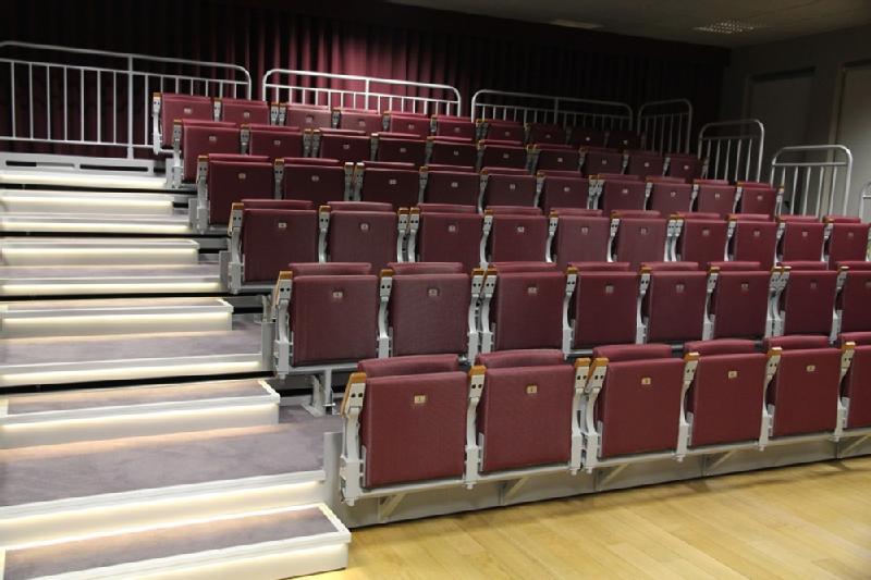 Sala wykładowa lub audytorium z rzędami pustych, bordowych siedzeń i oświetlonymi, wyłożonymi dywanem schodami po lewej stronie, oddzielonymi metalowymi poręczami.