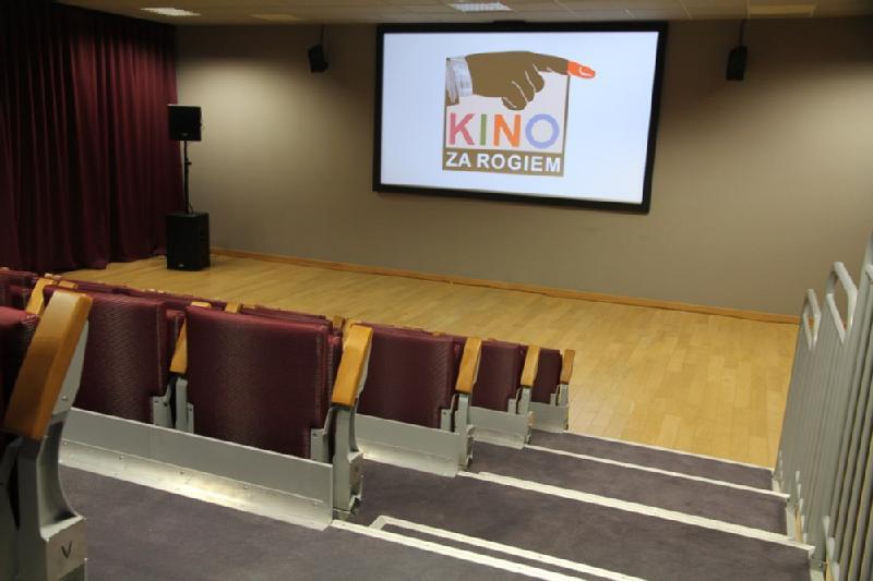 Małe, puste kino z czerwonymi siedzeniami, drewnianą podłogą i ekranem wyświetlającym logo „Kino Za Rogiem” ze wskazującą ręką.