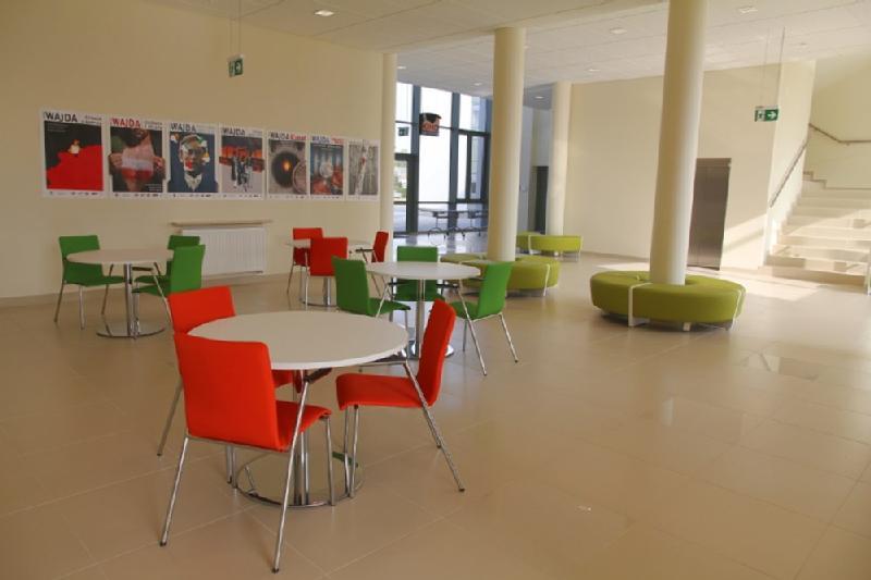 Nowoczesne, przestronne pomieszczenie z okrągłymi białymi stołami, czerwonymi i zielonymi krzesłami, okrągłymi zielonymi ławkami, dużymi oknami i plakatami filmowymi na ścianach. Przestrzeń wypełnia naturalne światło, a w tle widać schody.