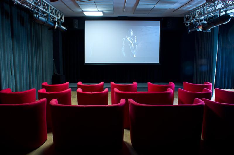 Mały, pusty teatr z rzędami jaskrawoczerwonych foteli zwróconych w stronę dużego ekranu wyświetlającego czarno-biały film. Pomieszczenie jest słabo oświetlone, ściany zasłaniają ciemne zasłony, a sufit jest oświetlony światłami scenicznymi.