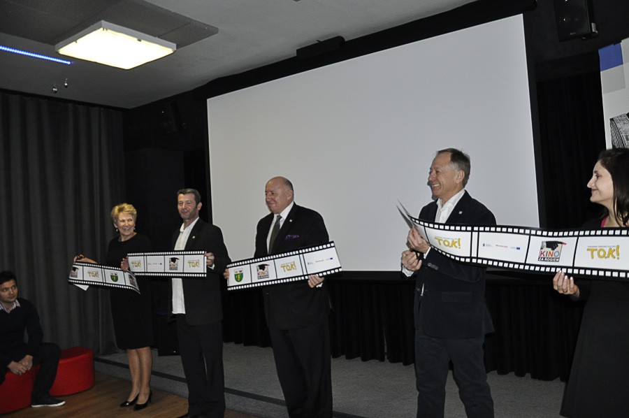 Pięć osób w formalnych strojach stoi przed dużym ekranem, uśmiechając się i trzymając długi, ozdobny baner z paskami filmowymi, jakby przecinały wstęgę na jakimś wydarzeniu. W tle widać ciemne zasłony i siedzącą osobę po lewej stronie.