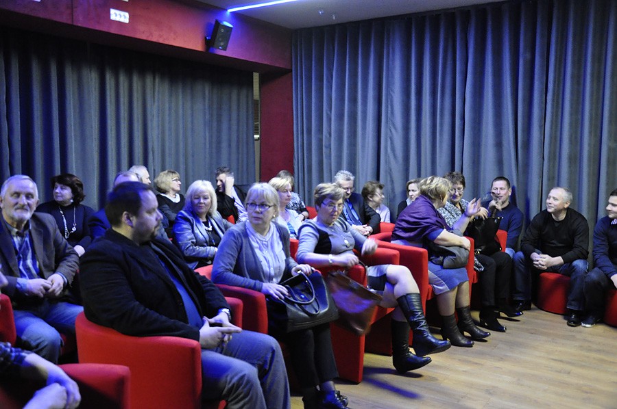 Grupa dorosłych siedzi na czerwonych krzesłach w słabo oświetlonym pokoju z szarymi zasłonami. Wyglądają, jakby czekali na rozpoczęcie wydarzenia, rozmawiając w małych grupach i wyglądając na zrelaksowanych.