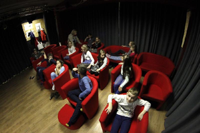 Dzieci siedzą i relaksują się w jaskrawoczerwonych fotelach ustawionych w rzędach w słabo oświetlonym pokoju z ciemnymi zasłonami i drewnianymi podłogami. Niektóre dzieci rozmawiają, podczas gdy inne patrzą przed siebie, tworząc zrelaksowaną, nieformalną atmosferę.