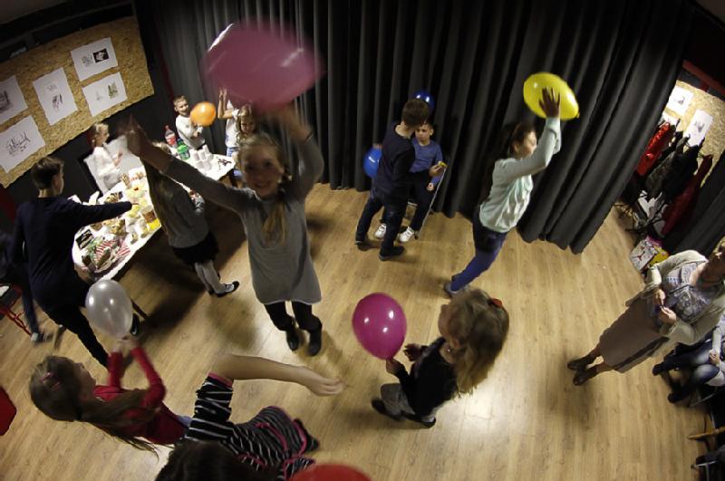 Dzieci bawią się kolorowymi balonami w pokoju z drewnianymi podłogami, podczas gdy grupa siedzi przy stole z jedzeniem i napojami z boku. Ciemne zasłony zasłaniają część tła. Scena jest żywa i radosna.