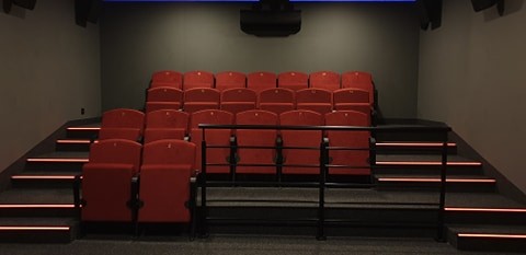 Mały teatr z trzema rzędami czerwonych siedzeń, słabym oświetleniem, czarnymi poręczami i oświetlonymi schodami prowadzącymi po obu stronach strefy siedzącej. Ekran nie jest widoczny.