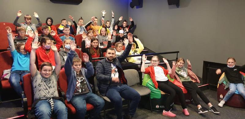 Grupa dzieci i jeden dorosły, niektórzy w maskach, siedzą na czerwonych i czarnych siedzeniach w sali o szarych ścianach i podnoszą ręce, uśmiechając się.