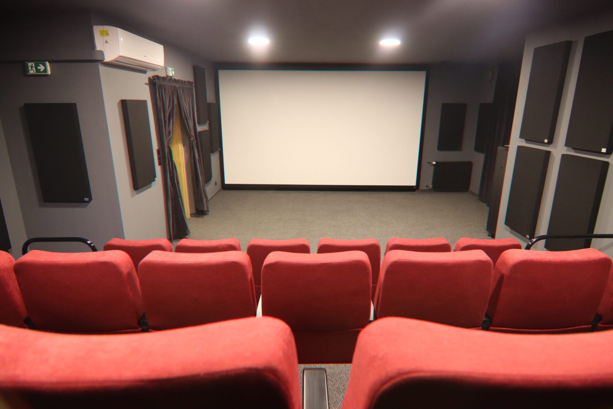 Mały pokój kina domowego z czerwonymi, wyściełanymi siedzeniami zwróconymi w stronę dużego, pustego ekranu, otoczony panelami akustycznymi na szarych ścianach pod oświetleniem sufitowym.