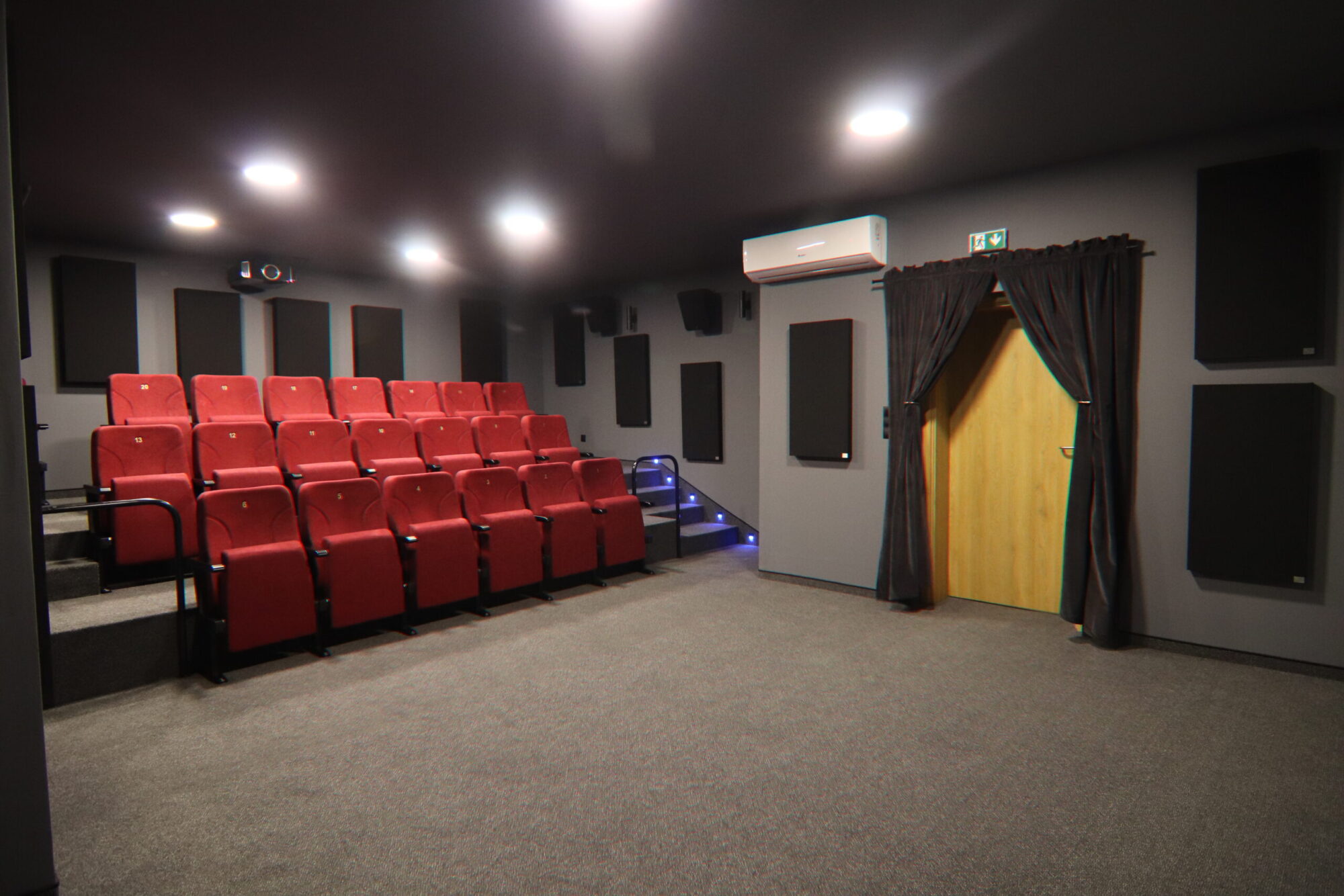 Niewielka sala kinowa z czerwonymi, wyściełanymi siedzeniami ustawionymi w rzędach naprzeciwko ekranu, szarymi ścianami z czarnymi panelami akustycznymi, drewnianymi drzwiami z ciemną zasłoną i oświetleniem sufitowym tworzącym przytulną atmosferę.