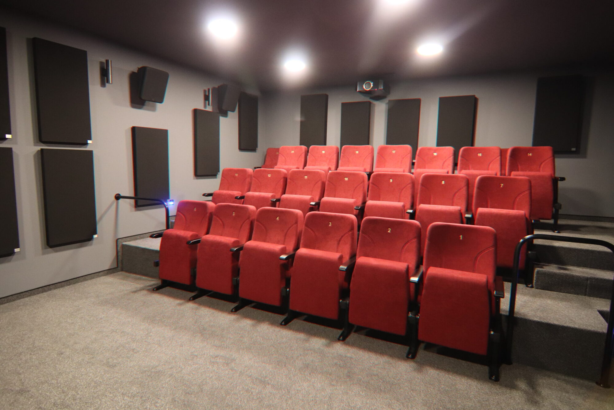Mały prywatny pokój kina domowego z rzędami czerwonych, wyściełanych siedzeń, szarą wykładziną, czarnymi panelami akustycznymi na ścianach, głośnikami i projektorem zamontowanym na suficie. Pokój jest dobrze oświetlony lampami sufitowymi.