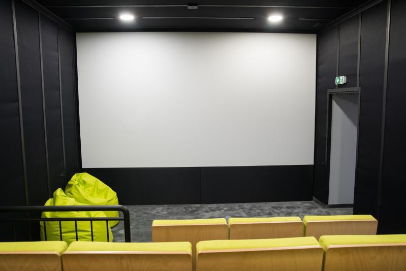 Małe, puste kino z żółtymi siedzeniami, dużym, pustym ekranem, ciemnymi ścianami i pojedynczym zielonym krzesłem-workiem z fasolą przy wejściu, obok drzwi wyjściowych.
