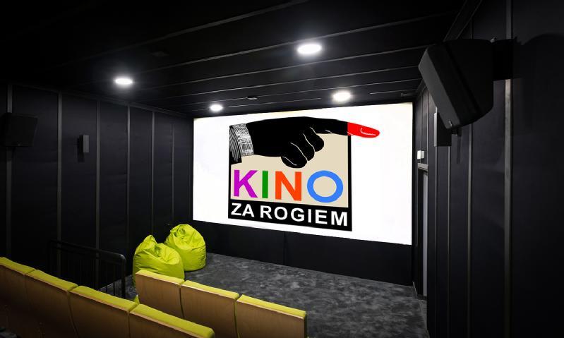 Małe, nowoczesne kino z żółtymi siedzeniami, zielonymi workami wypełnionymi fasolą i dużym ekranem wyświetlającym logo z wskazującą ręką i napisem KINO ZA ROGIEM wykonanym kolorowymi literami.