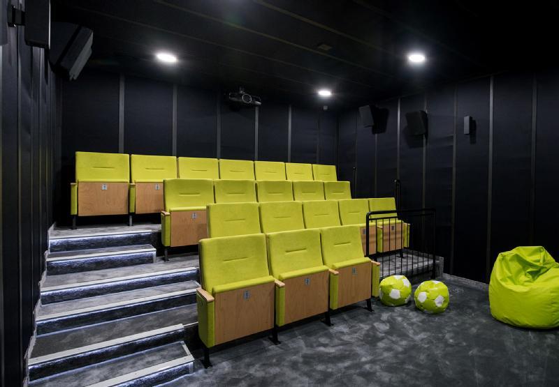 Mała, nowoczesna sala teatralna z żółtymi siedzeniami ustawionymi w rzędach, czarnymi ścianami, szarym dywanem oraz dwoma zielonymi workami sako i workiem w kształcie piłki nożnej z przodu po prawej stronie. Oświetlenie sufitowe oświetla przestrzeń.