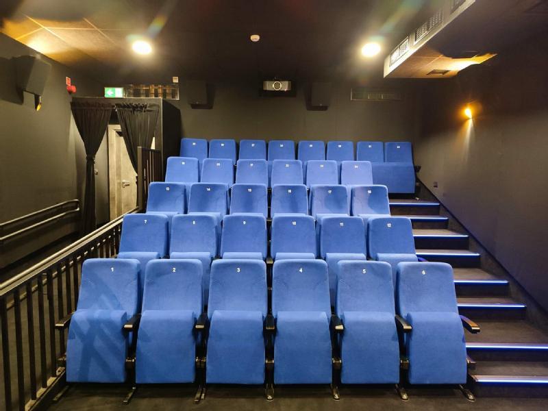 Niewielka sala kinowa z trzema rzędami ponumerowanych niebieskich siedzeń, czarnymi ścianami i schodami z oświetlonymi krawędziami po prawej stronie. Nad siedzeniami znajduje się projektor, a z tyłu po lewej stronie są drzwi wyjściowe.