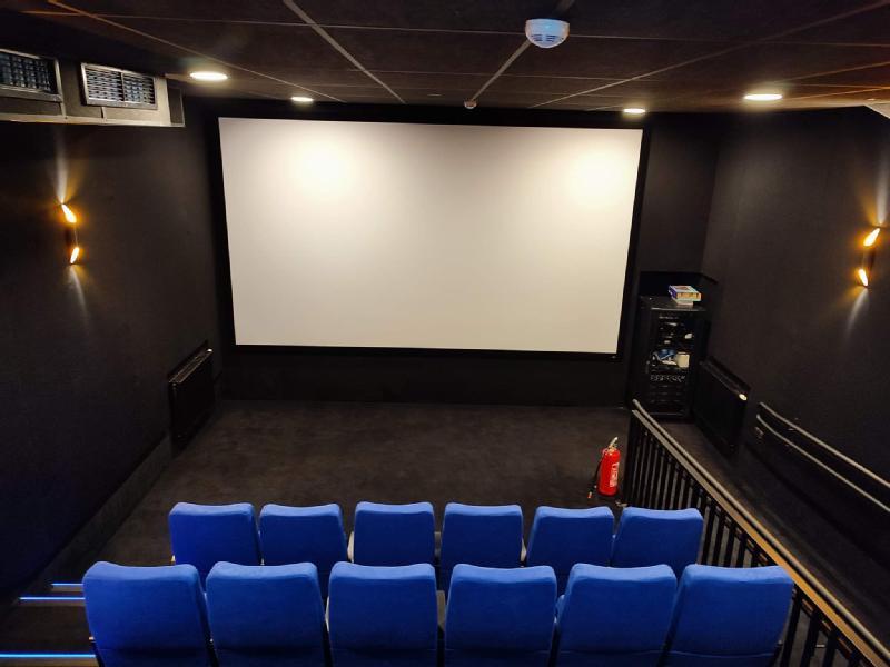 Małe, nowoczesne kino z niebieskimi siedzeniami, dużym pustym białym ekranem, czarnymi ścianami, kinkietami, gaśnicą i stojakiem na sprzęt wzdłuż boku. Pokój wydaje się pusty.