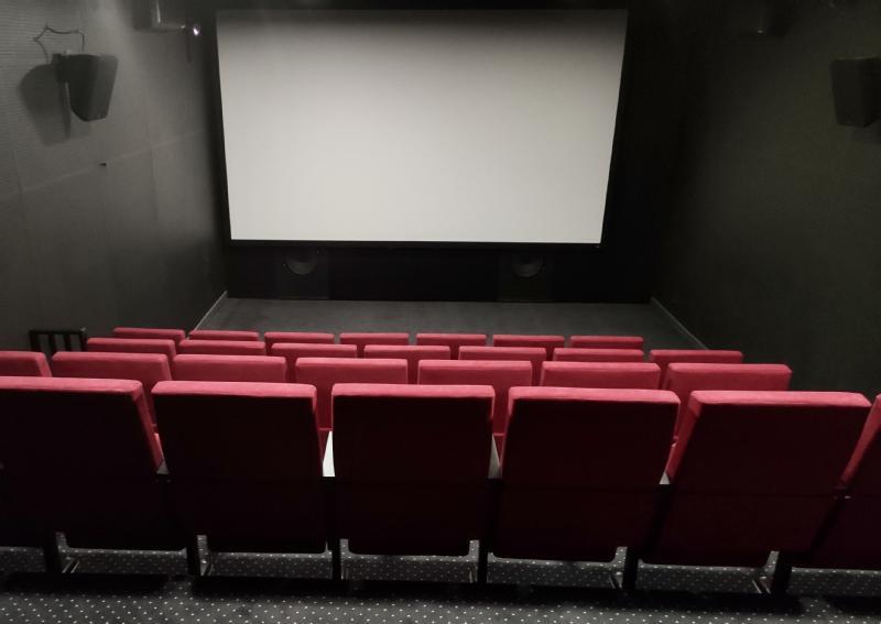 Puste kino z rzędami czerwonych siedzeń ustawionych naprzeciwko dużego, pustego ekranu, otoczone ciemnymi ścianami i dywanami w kropki.