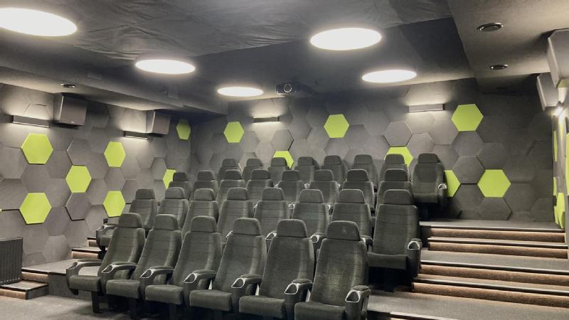 Małe, nowoczesne kino z piętrowymi siedzeniami i szarymi krzesłami. Ściany zdobią szare i zielone heksagonalne panele, a okrągłe lampy sufitowe jasno oświetlają pomieszczenie.