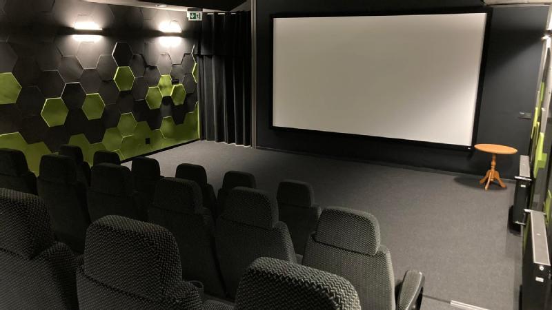 Małe, nowoczesne kino z czarnymi siedzeniami, dużym pustym ekranem, sześciokątnymi panelami ściennymi w kolorze czarnym i zielonym oraz okrągłym drewnianym stolikiem bocznym przy ekranie. Kino jest puste i dobrze oświetlone.