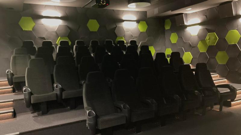 Mały, pusty teatr z rzędami czarnych siedzeń i ścianami ozdobionymi czarno-zielonymi sześciokątnymi wzorami, oświetlony kinkietami.