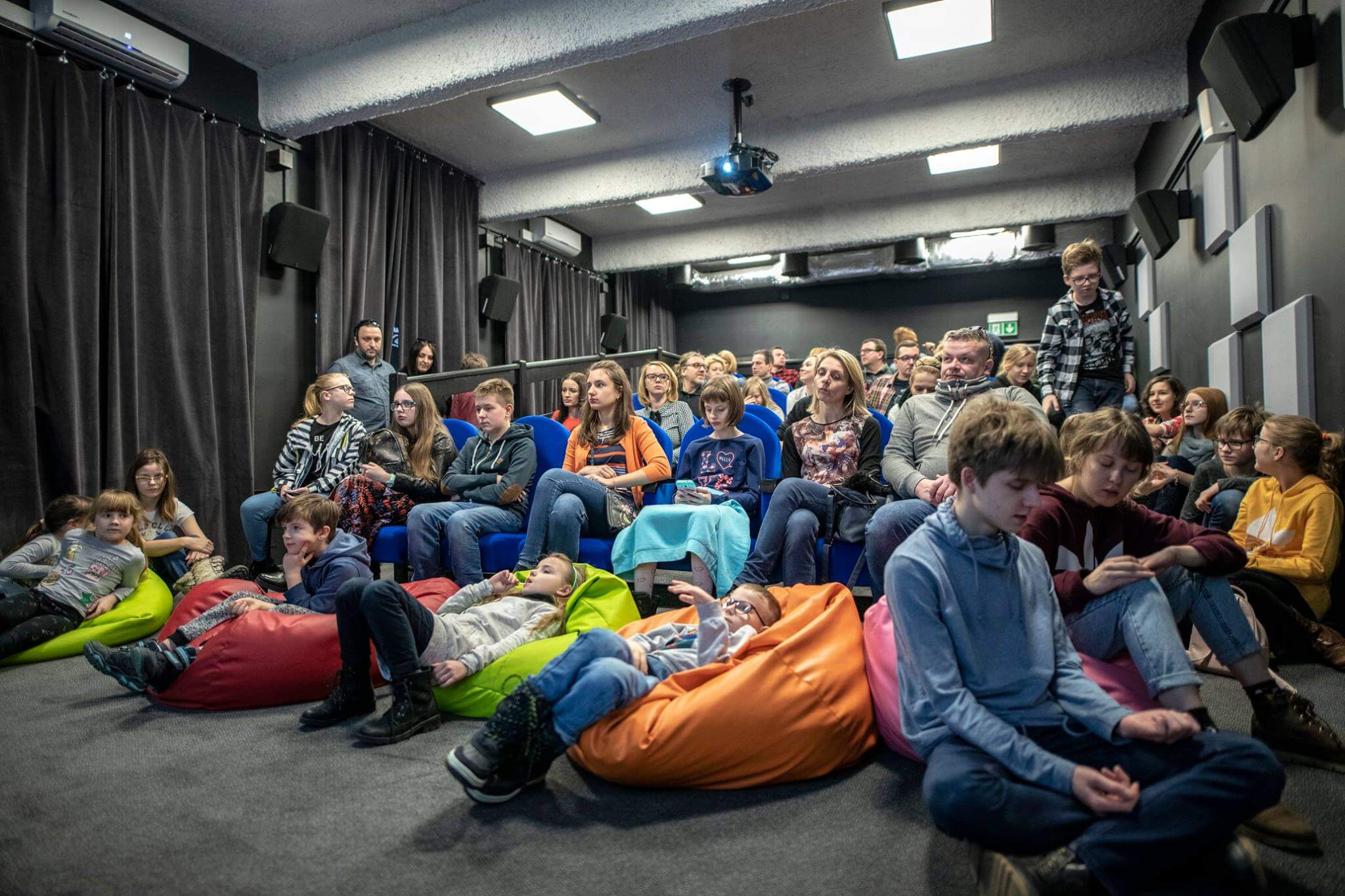 Grupa dzieci i dorosłych siedzi na workach fasolowych i krzesłach w małym teatrze lub klasie, uważnie obserwując coś poza ramką. W pomieszczeniu są ciemne ściany, zasłony i projektor sufitowy.