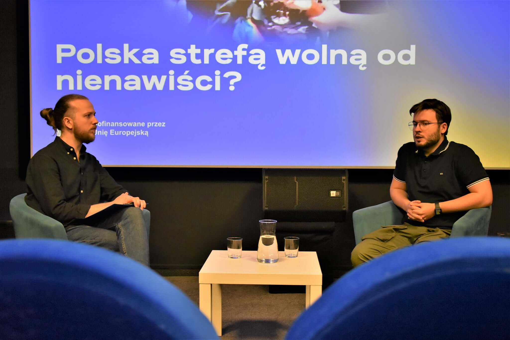 Dwóch mężczyzn siedzi naprzeciwko siebie na scenie z mikrofonami i prowadzi dyskusję. Za nimi znajduje się ekran wyświetlający tekst w języku polskim: Polska strefa obowiązana od nienawiści? Pomiędzy nimi znajduje się mały stolik ze szklankami z wodą.