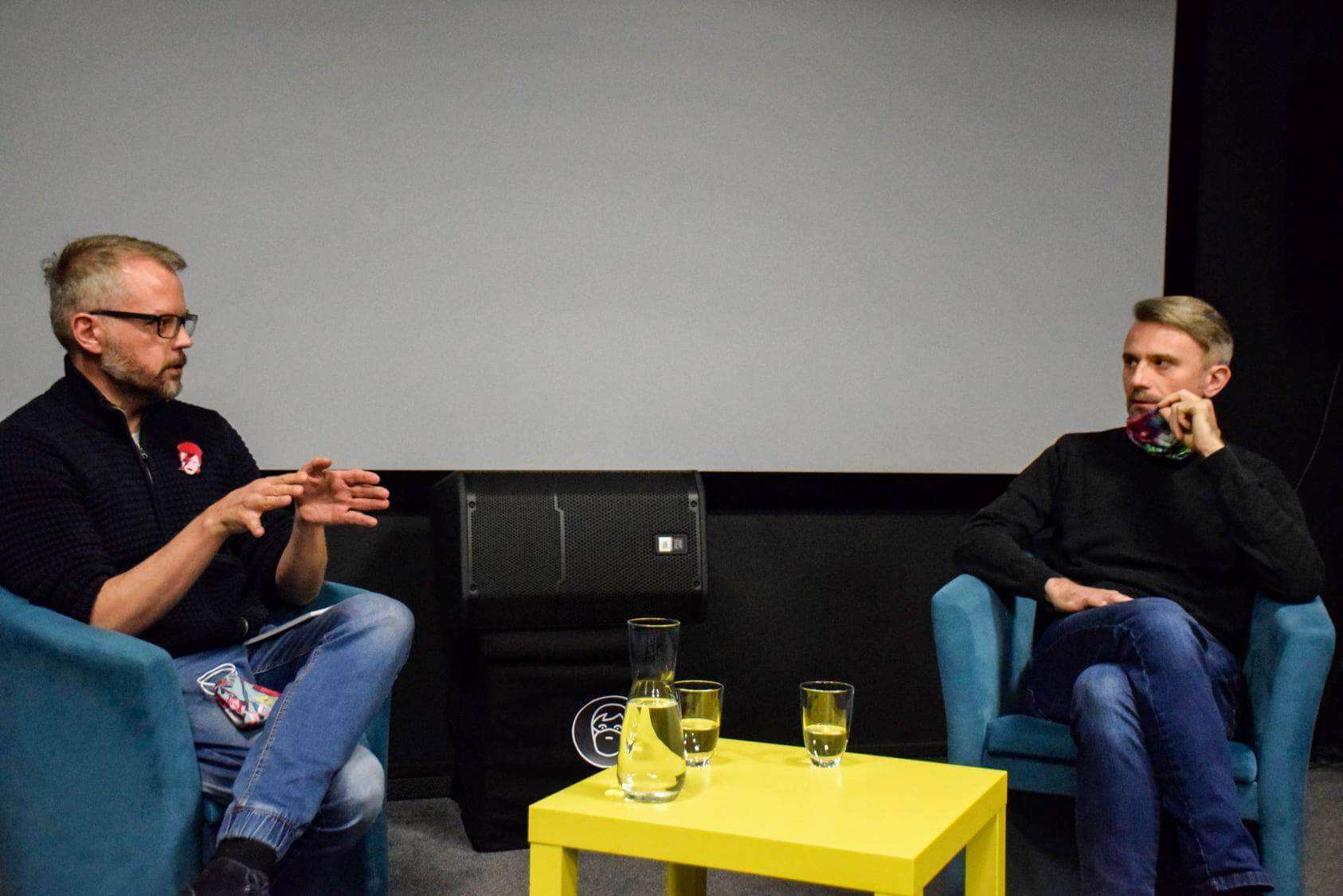 Dwóch mężczyzn siedzi na niebieskich fotelach naprzeciwko siebie, rozmawiając. Między nimi znajduje się mały żółty stolik z trzema szklankami wody. W tle widać duży ekran i głośnik.