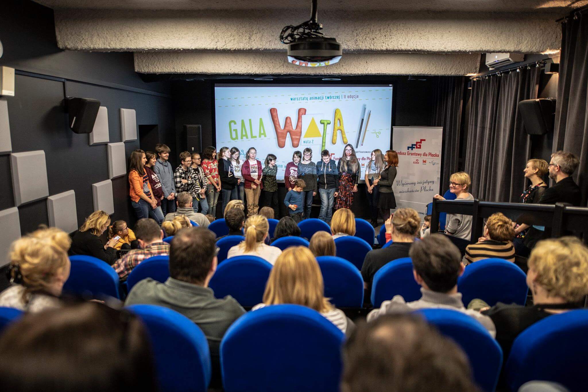 Grupa ludzi stoi na scenie przed ekranem wyświetlającym „Gala WATA”, podczas gdy publiczność na niebieskich krzesłach ogląda w małym teatrze. Kobieta z mikrofonem zwraca się do publiczności.