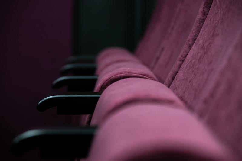 Zbliżenie na kilka bordowych aksamitnych foteli teatralnych ustawionych w rzędzie, ukazujące podłokietniki i oparcia siedzeń na ciemnym, rozmytym tle.