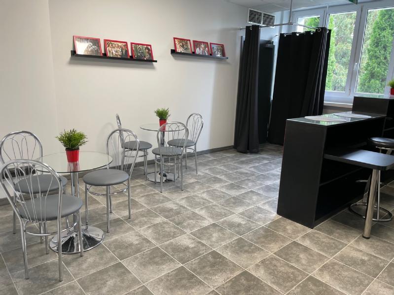 Mały, nowoczesny pokój ze szklanymi stołami i metalowymi krzesłami, każdy stół ozdobiony rośliną doniczkową. Czarne zasłony zasłaniają część okna, a oprawione zdjęcia stoją na półce nad stołami.