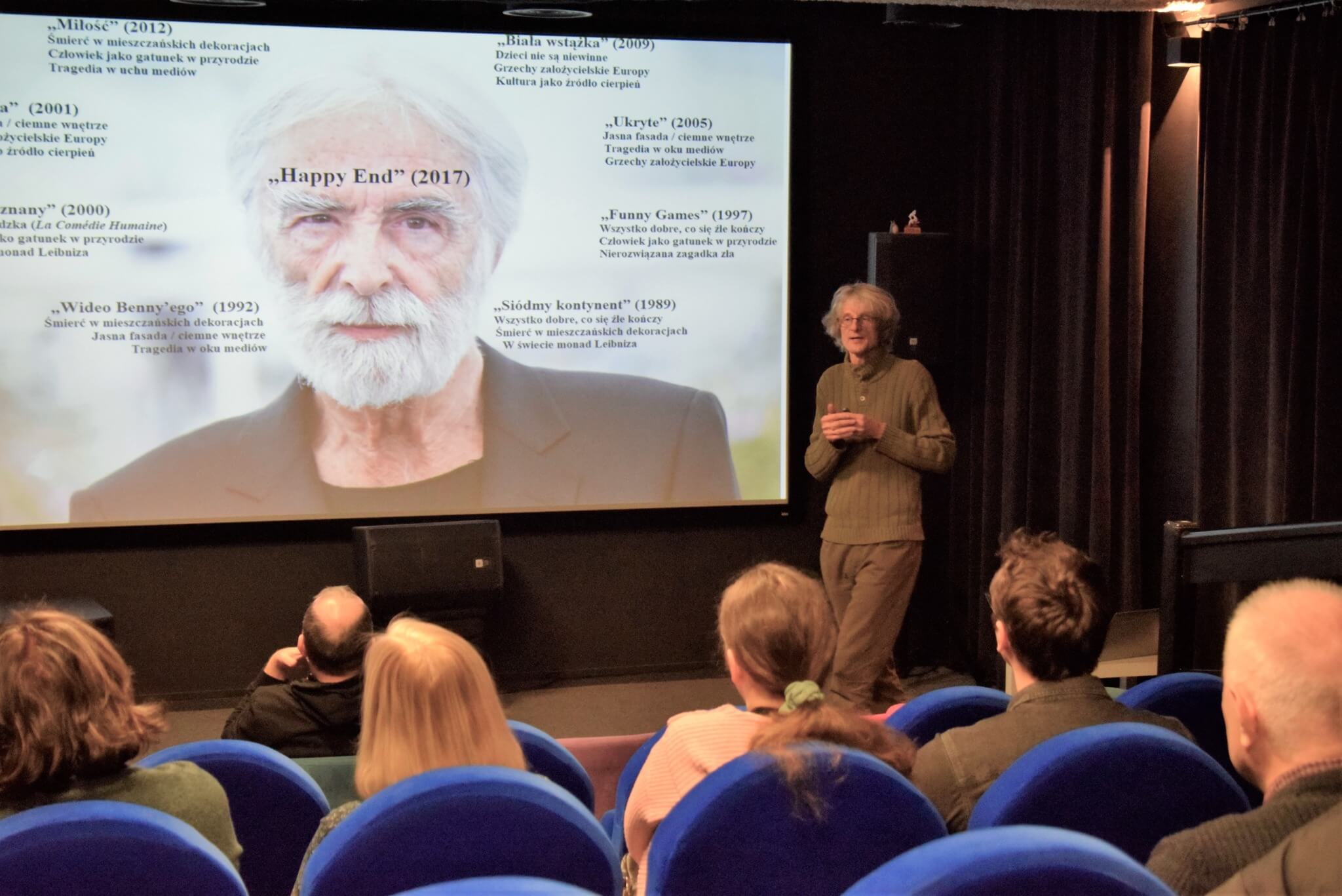 Osoba stoi przed dużym ekranem wyświetlającym informacje o filmach, zwracając się do publiczności siedzącej na niebieskich krzesłach w małym teatrze. Na ekranie znajduje się portret i tekst w języku polskim.