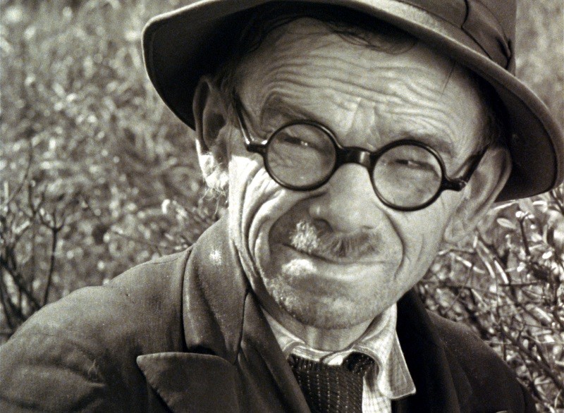 Starszy mężczyzna w okrągłych okularach, kapeluszu fedora i marynarce z krawatem mruży oczy i lekko się uśmiecha, patrząc w kamerę. W tle widać krzewy i liście.
