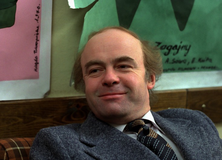 Mężczyzna w średnim wieku z przerzedzonymi włosami i delikatnym uśmiechem, ubrany w szary garnitur i krawat w paski, siedzi w pomieszczeniu przed kolorowymi plakatami na ścianie.