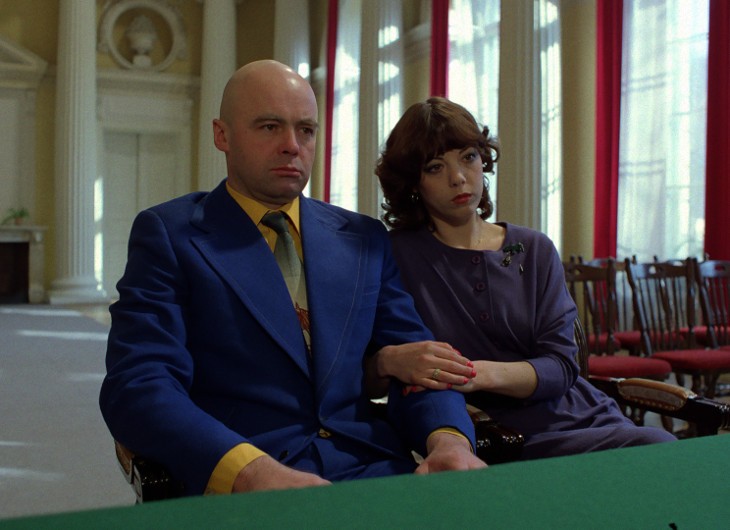 Mężczyzna w niebieskim garniturze i kobieta w fioletowej sukience siedzą blisko siebie w ogromnym pomieszczeniu z wysokimi kolumnami i czerwonymi zasłonami. Oboje wyglądają poważnie i poważnie.