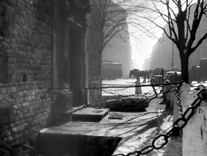 Czarno-białe zdjęcie miejskiej ulicy z wozem konnym, kocimi łbami, gołymi drzewami i łańcuchami wzdłuż chodnika. Wczesne poranne światło rzuca długie cienie. W tle widać tramwaj.