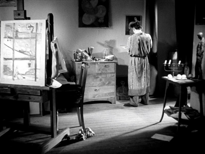 Osoba w długiej szacie stoi naprzeciwko komody w pracowni artystycznej, otoczona obrazami, rysunkami i artykułami artystycznymi. Scena jest oświetlona dramatycznymi cieniami, co daje kontemplacyjną, kreatywną atmosferę.